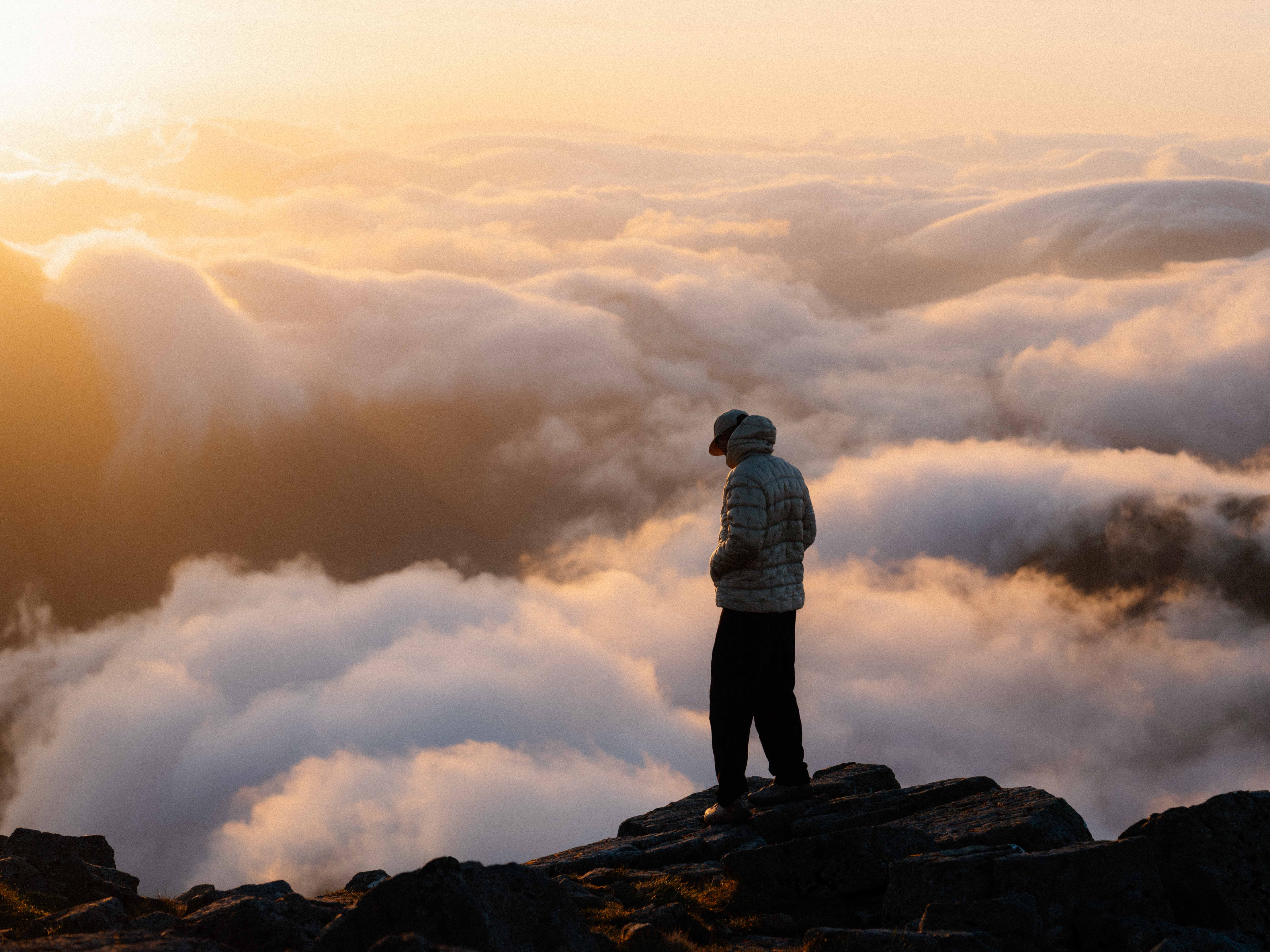 A man stands on the edge of a cliff looking down at the clouds